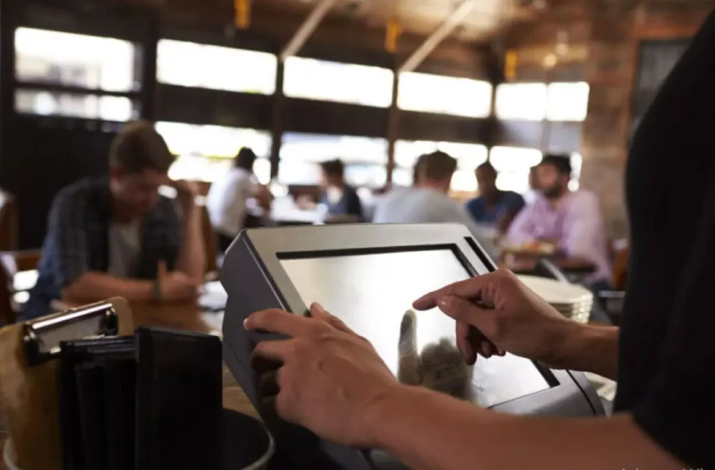 Atendente utilizando um sistema para restaurante em um tablet. Ao fundo, pessoas estão sentadas ao redor de diversas mesas.