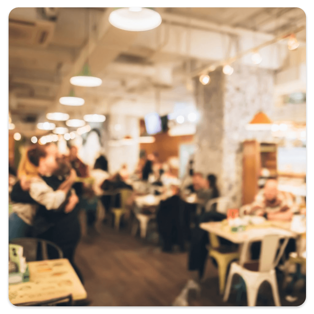 Imagem desfocada de um salão de restaurante, mostrando várias mesas e pessoas ao fundo.